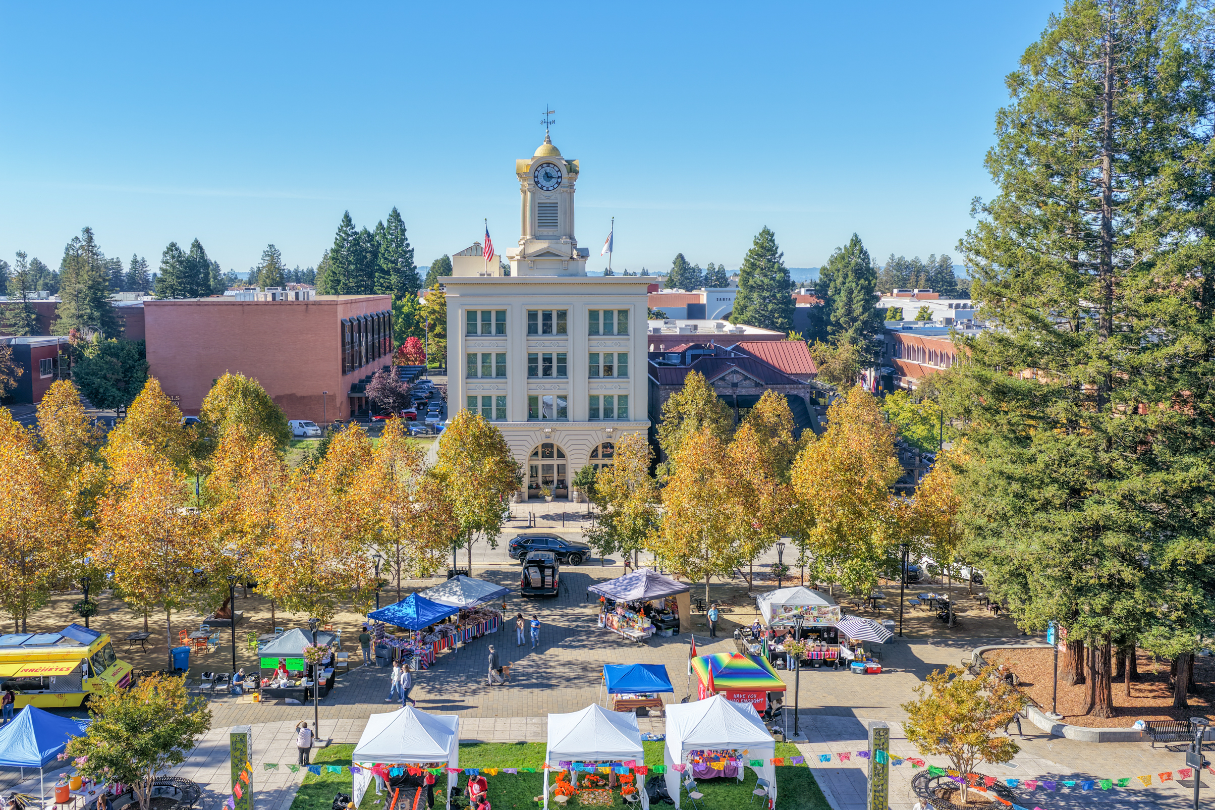 Aerial view of a town square featuring a clock tower building surrounded by trees with autumn foliage and various vendor tents.