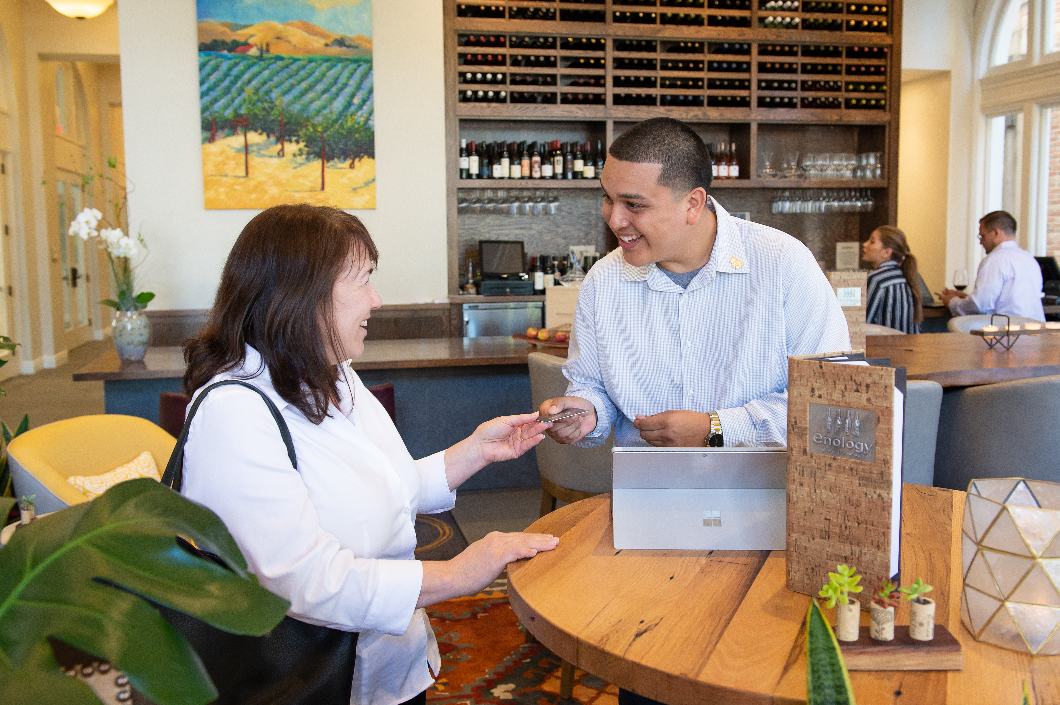 A cheerful interaction at a hospitality venue, with a staff member assisting a guest at the reception desk, surrounded by a modern interior featuring a wine display and decorative plants.